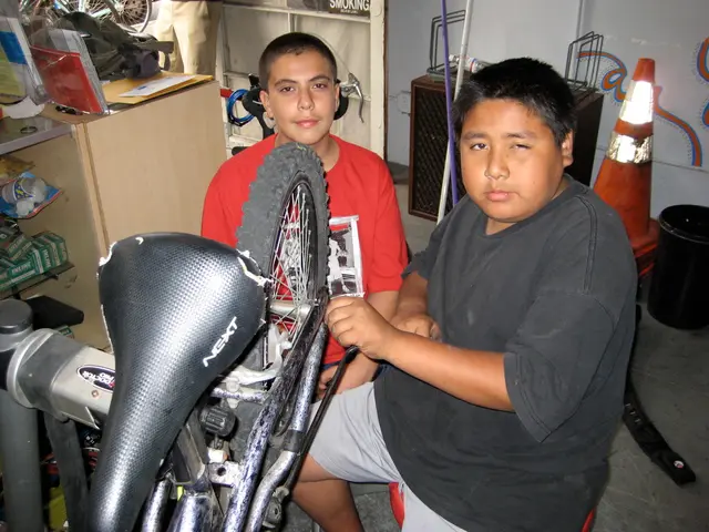 In the center of the picture there are two people repairing a bicycle. On the left there is desk,...
